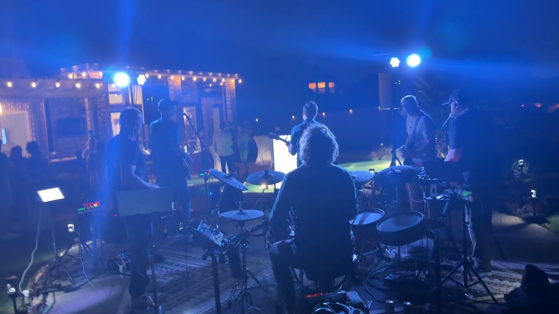 View from behind the drummer, looking past the band to the backyard crowd under blue stage lights.