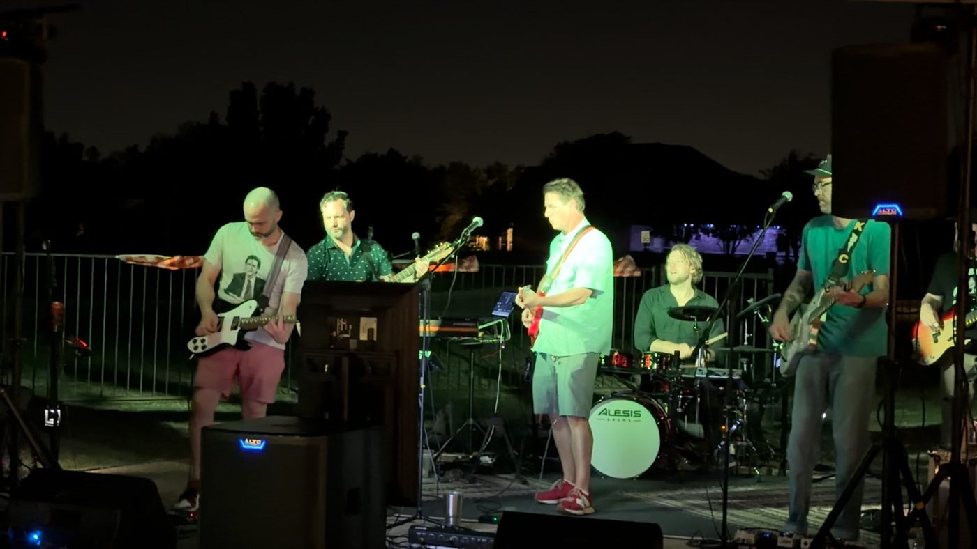 The whole band on a backyard stage at night: guitars across the front, drummer behind.
